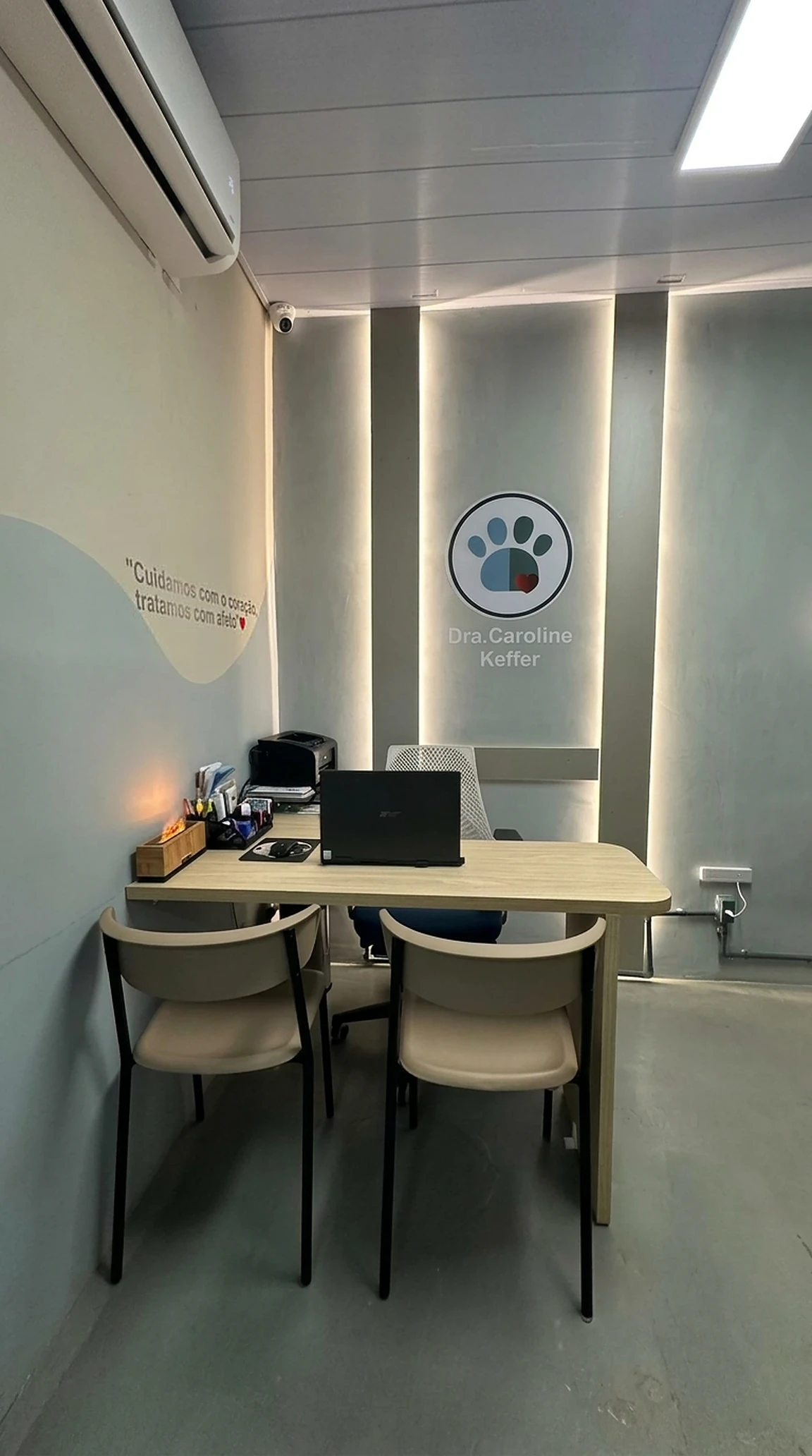 Consultório da clínica visto de frente, com a mesa de atendimento, duas cadeiras, o selo da Dra. Caroline Keffer iluminado na parede e a frase "Cuidamos com o coração, tratamos com afeto"